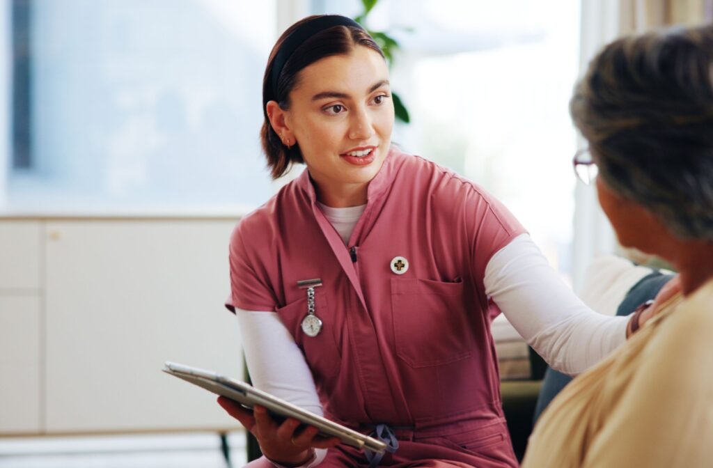 A caregiver in senior living touches an older adult's arm and reassures them while holding a clipboard during a regular check-in