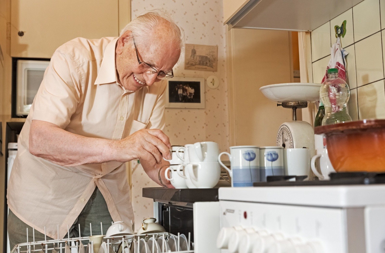 A smiling senior in independent living empties a dishwasher and stacks teacups on top of the counter.