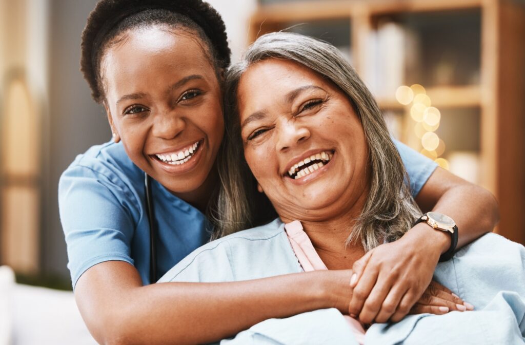 A caregiver hugs a senior resident from behind as they both smile toward the camera.