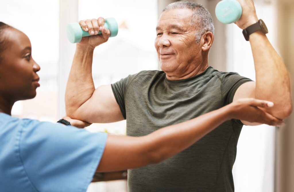 A senior lifts light dumbbells with both arms as a caregiver assists with correcting their form.